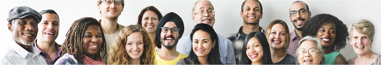 A diverse group of people of various ages, genders, and ethnicities stand close together, smiling warmly at the camera against a plain white background.